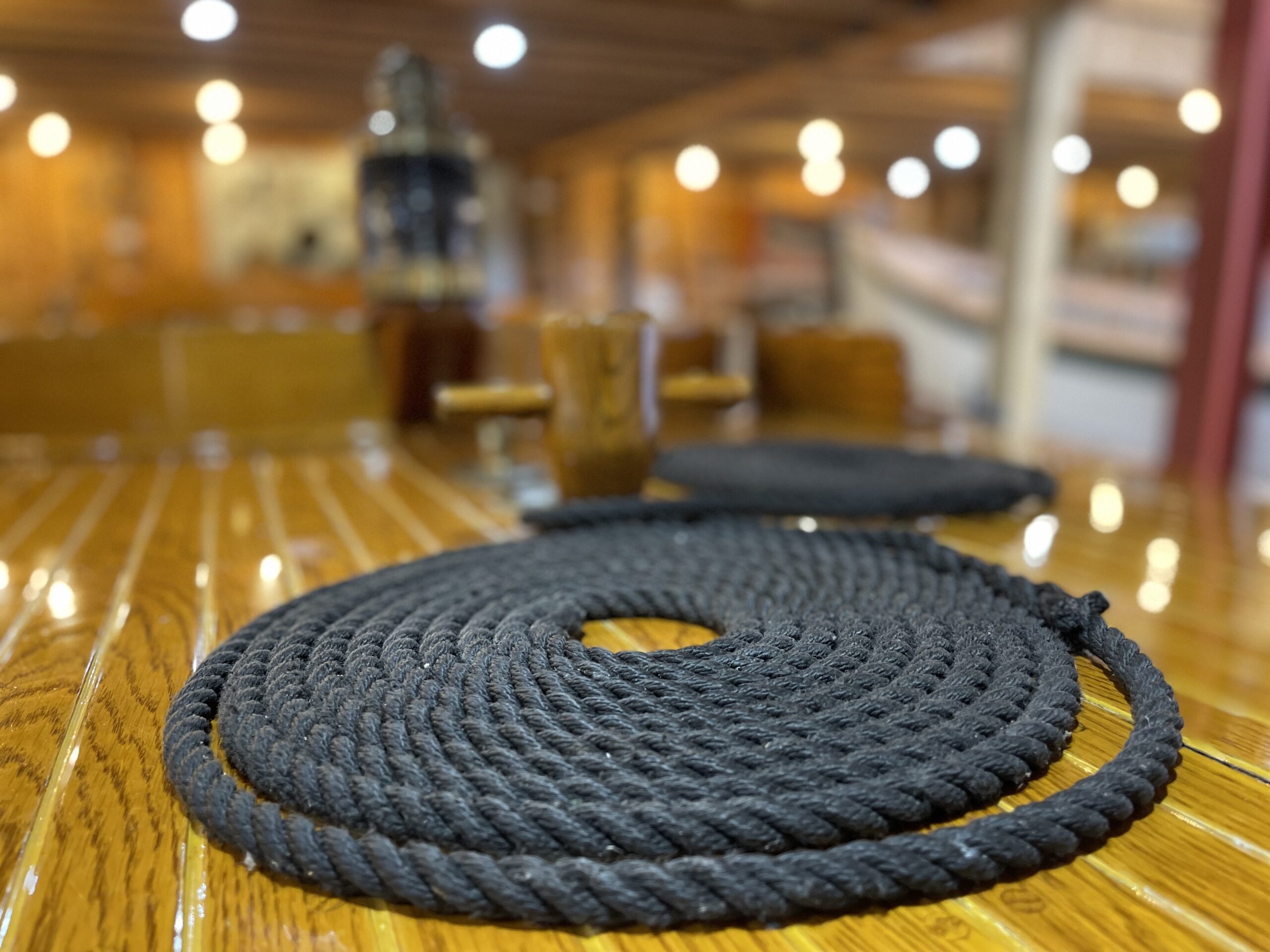 A neatly coiled black rope on a polished wooden table, with blurred lights and maritime equipment in the background.