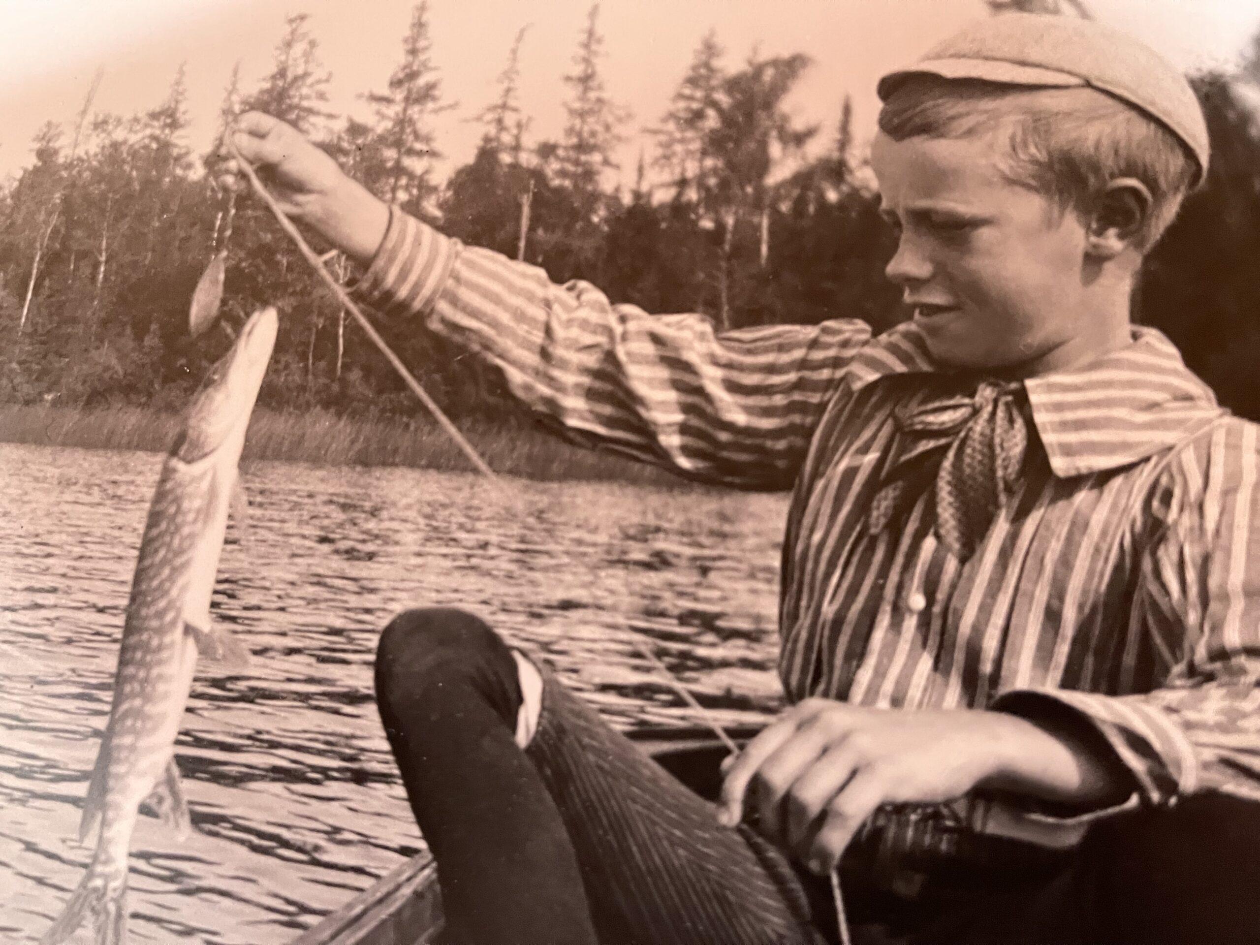 A vintage photo of a young boy in a striped shirt and cap, holding up a freshly caught fish on a line, sitting in a boat on a lake.