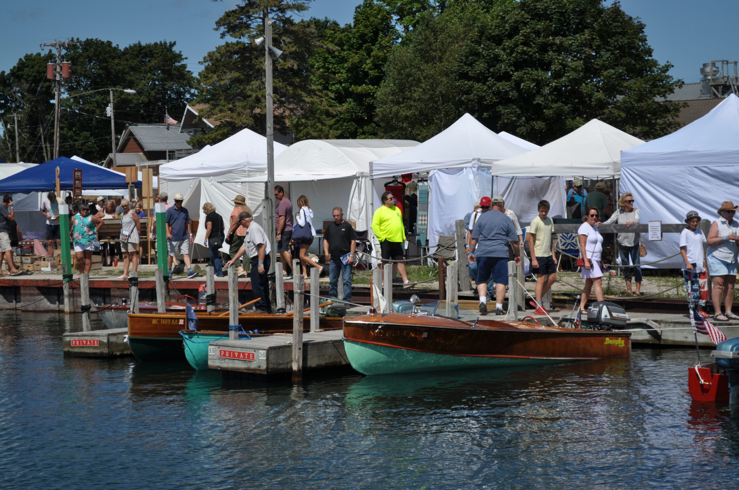 People and tents near docked wooden boats at a sunny outdoor event.