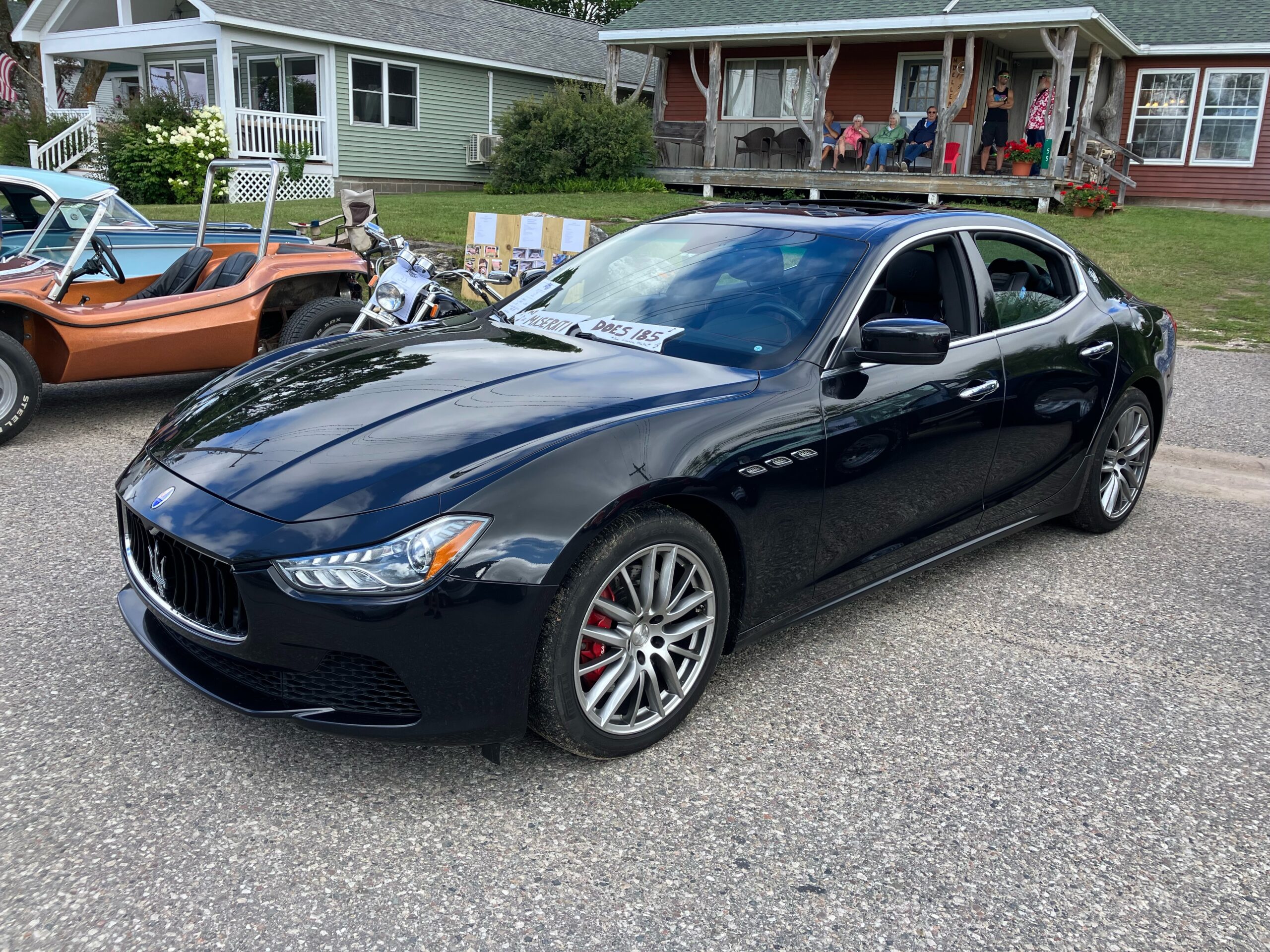 Shiny black Maserati parked at an outdoor event, with other classic cars and motorcycles in the background.