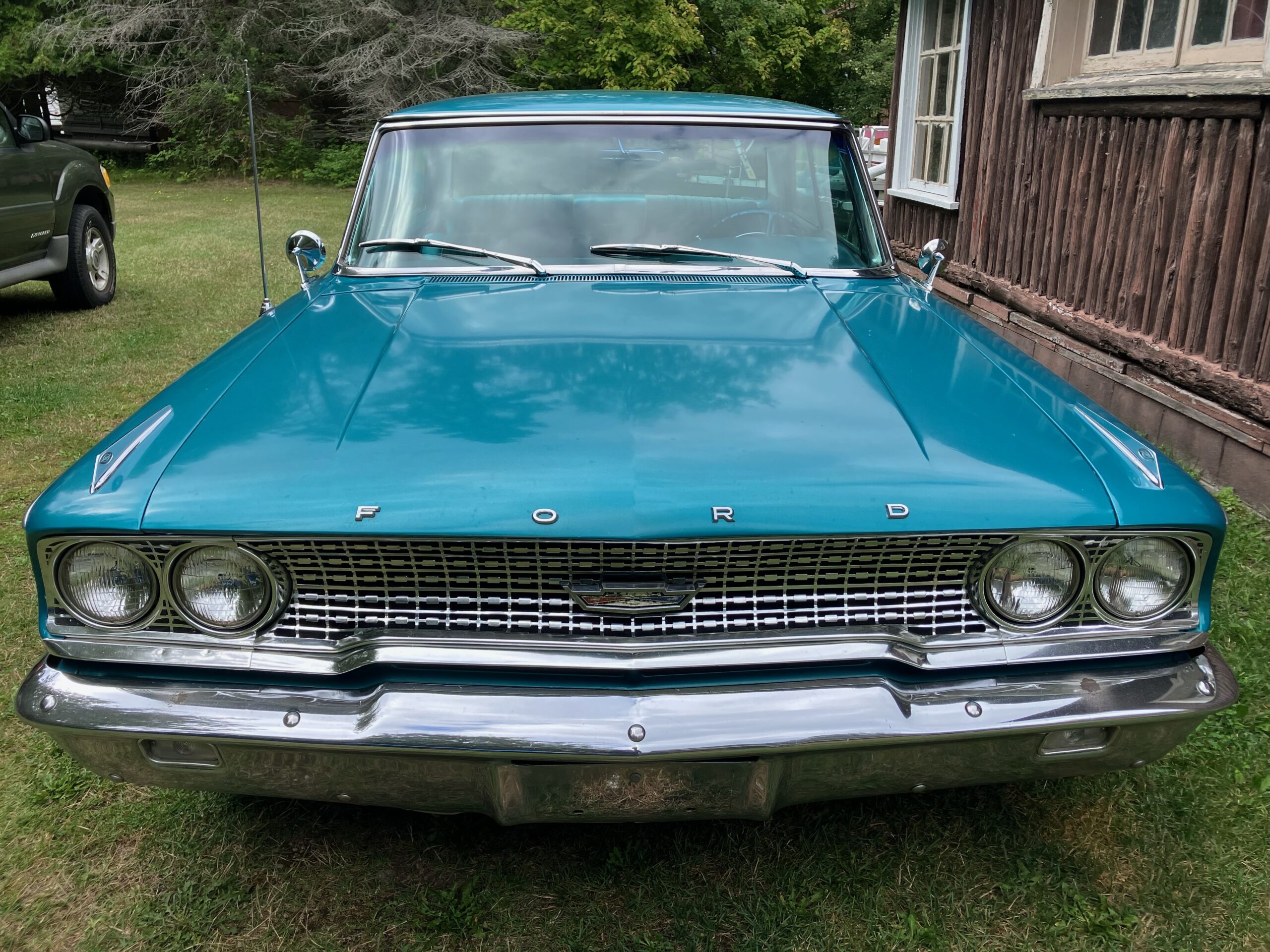 A teal vintage Ford car parked on grass next to a rustic wooden building.