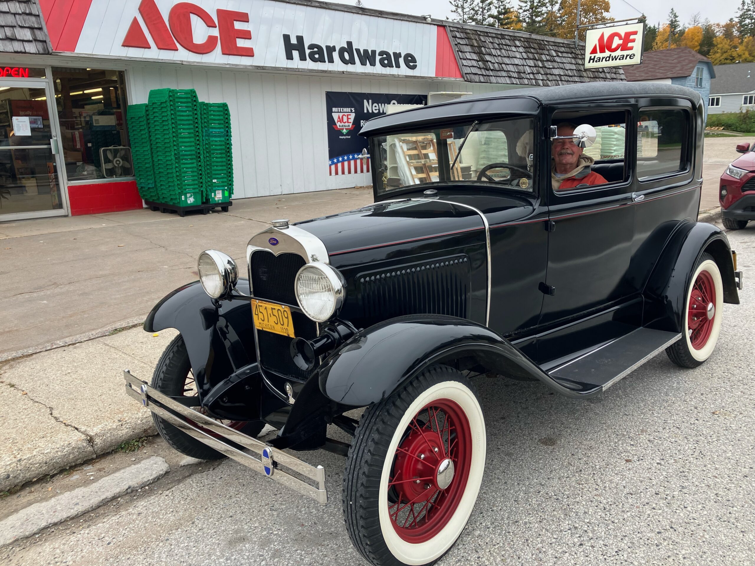 A man smiling from the driver's seat of a black vintage car with red wheels, parked in front of an ACE Hardware store.