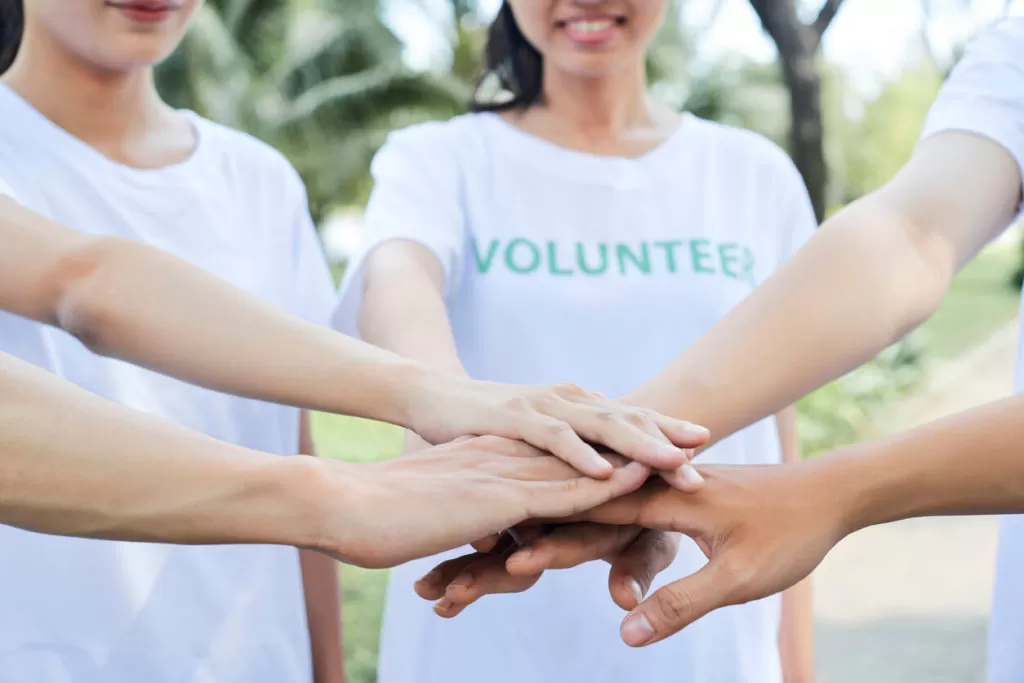 A group of volunteers wearing white shirts, placing their hands together in a stack, symbolizing teamwork.